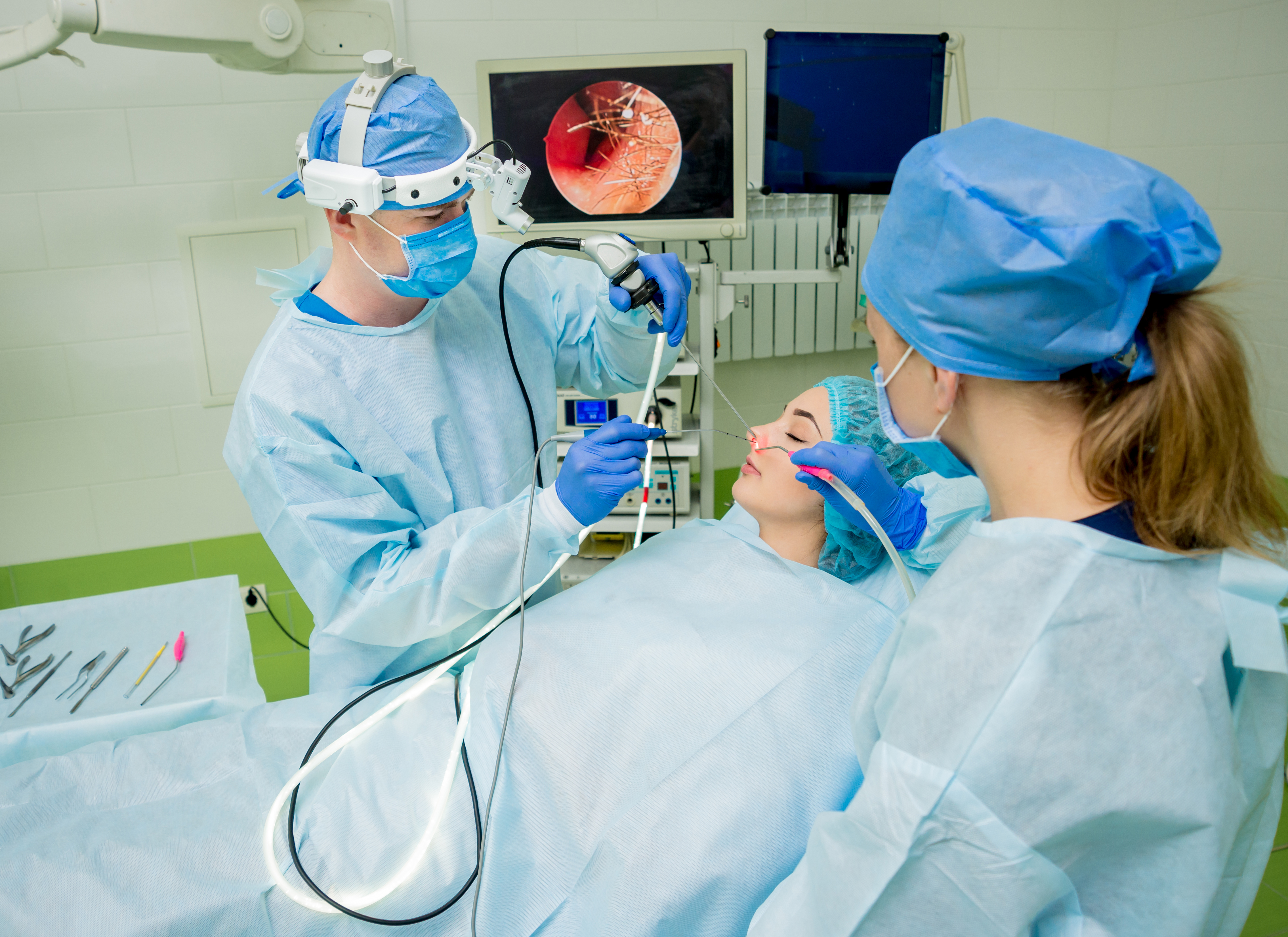 Two medical professionals in full surgical PPE performing endoscopic sinus surgery on a patient.