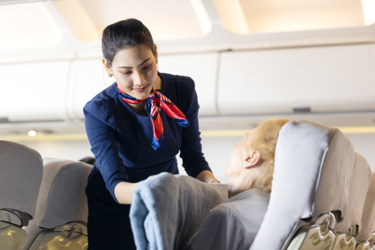 Photograph of an airhostess handing a blanket to a passenger who is sitting in their seat and smiling.
