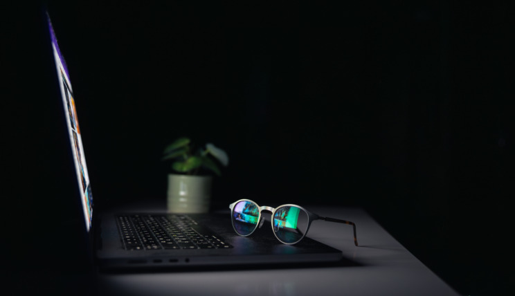Photo of a pair of glasses sat on the keypad of a laptop in a dimly lit room.