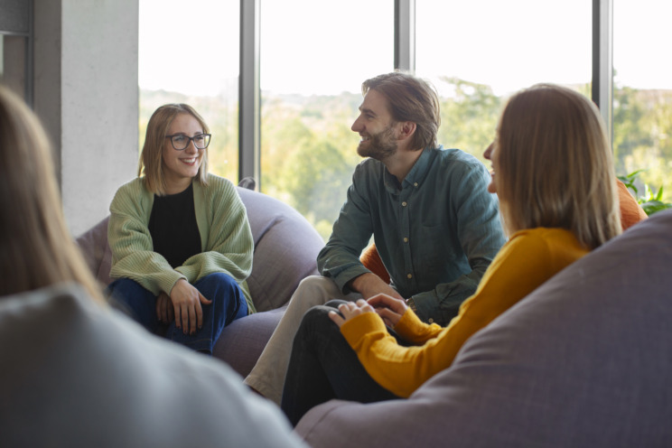 Photograph of three people sat on sofas smiling and looking in different directions as if in a larger group outside of the photo. One person wears glasses.