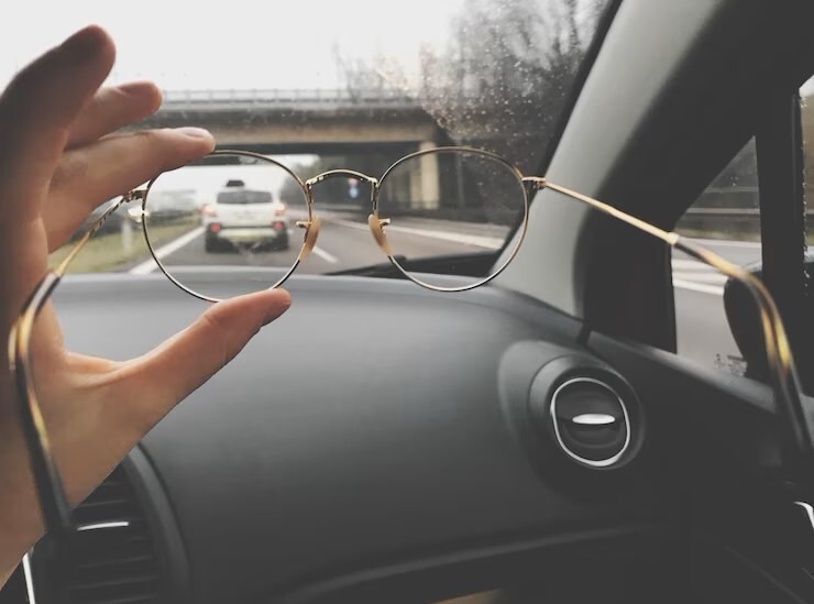 Photograph of a hand holding up glasses in front of the view out of the front of a car.