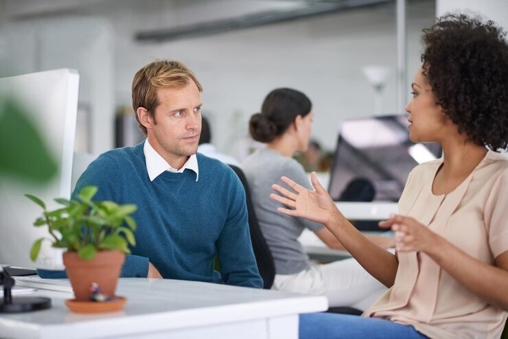 Photo of two people having a conversation at a desk, using their hands expressively.