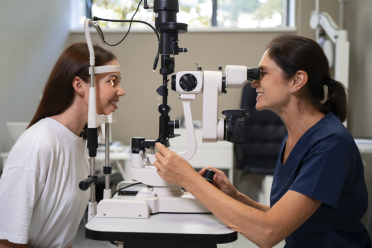 Photo of someone with their head rested in the head rest of an autorefractor and an ophthalmologist sat opposite them, looking into the eyepiece. Both people are smiling.