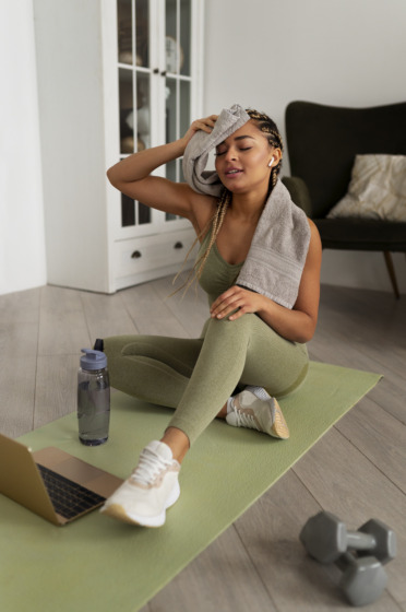 Photograph of a person sat on a yoga mat wiping their forehead with a towel with dumbbells next to them.